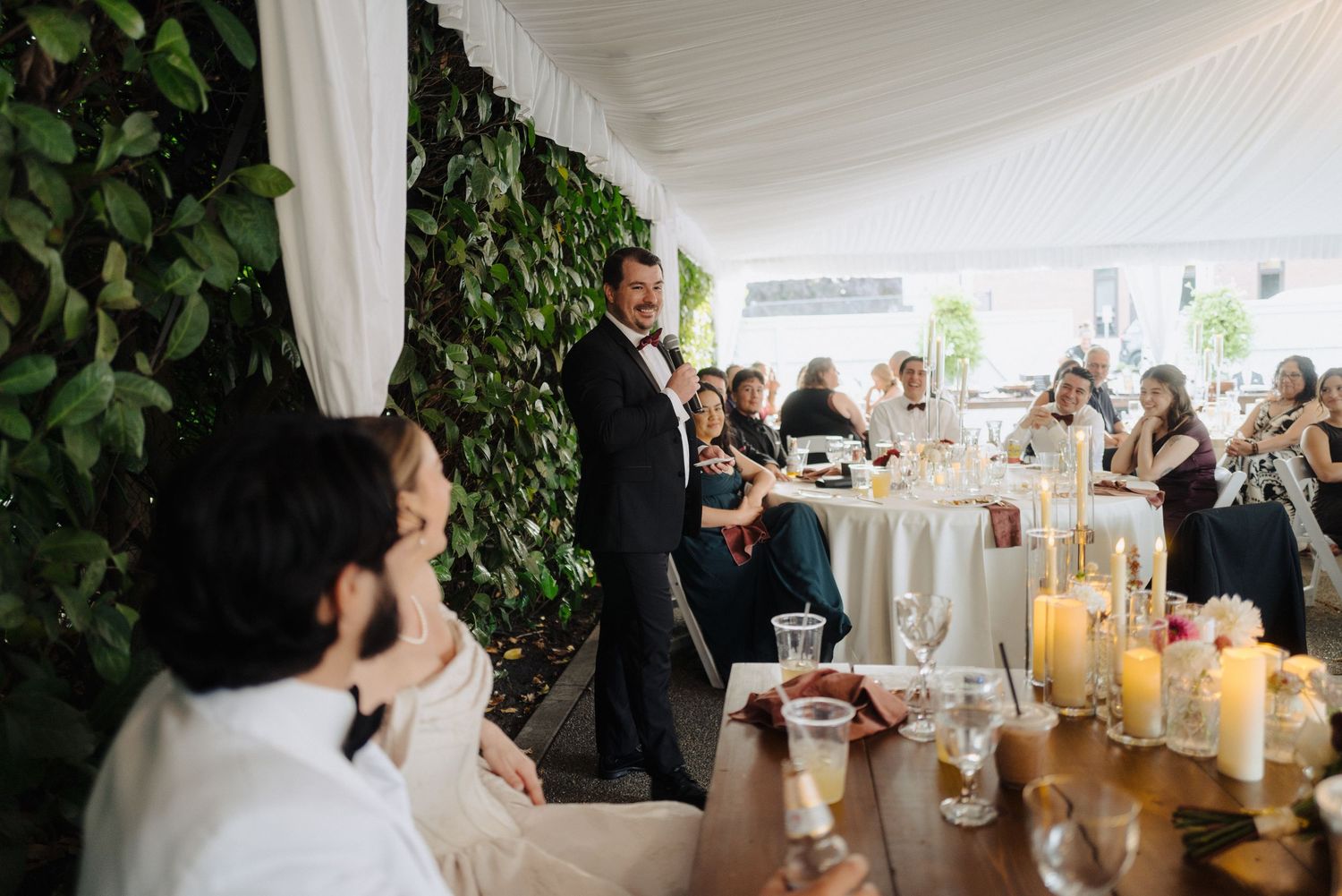 Wedding reception scene with guests seated at candlelit tables decorated with greenery.