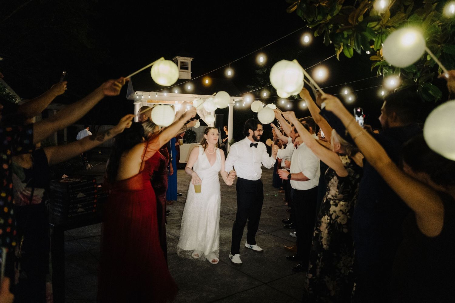 Wedding guests dance and celebrate on an outdoor patio decorated with glowing paper lanterns at night.