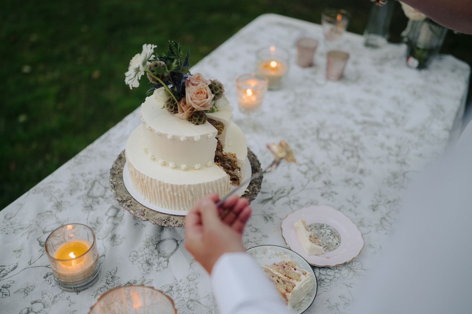 Wedding cake and candles displayed on a white lace tablecloth at an outdoor reception.