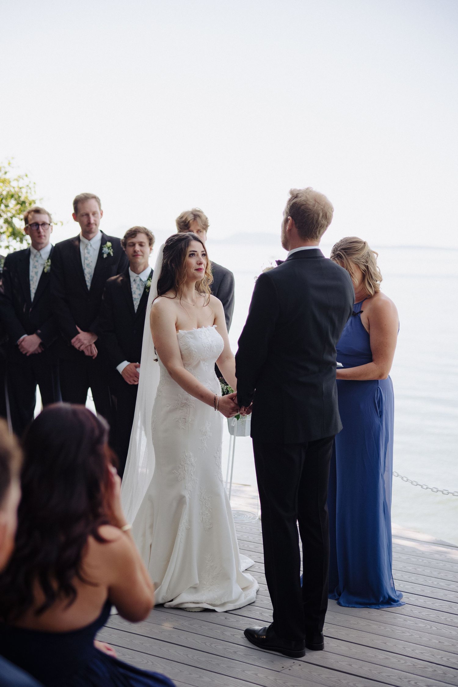 Intimate waterfront wedding ceremony with attendants in formal attire gathered on a wooden deck.