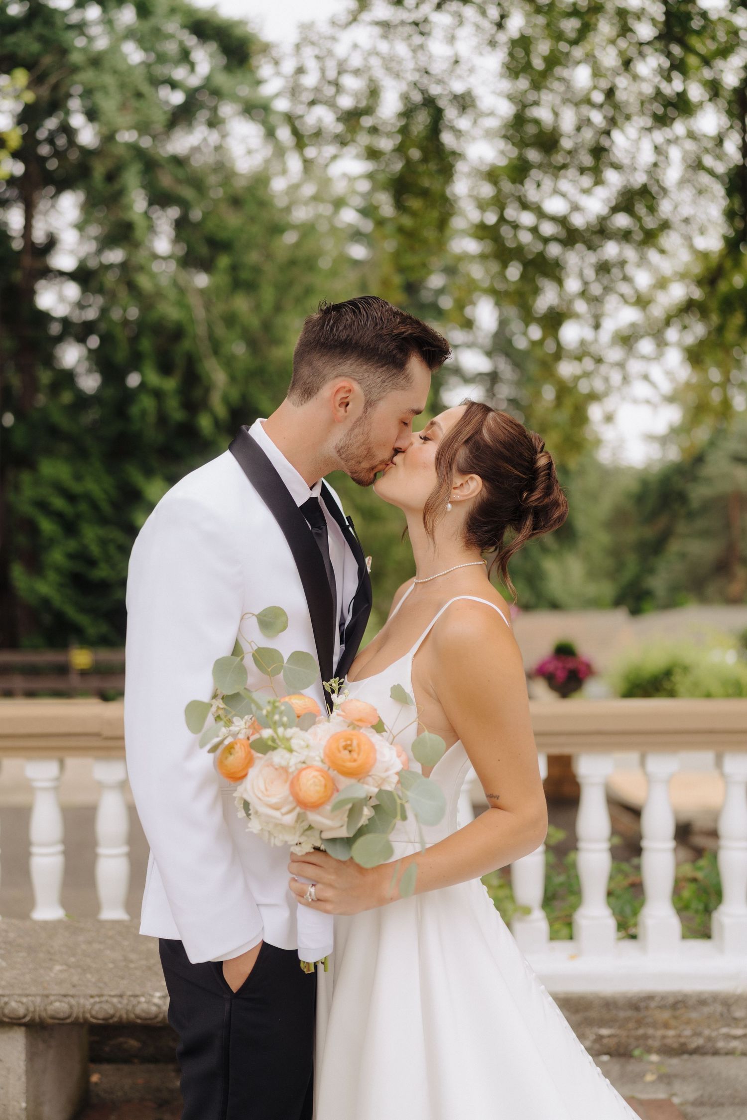 Romantic wedding couple embracing by white balustrade with peach and eucalyptus bouquet.