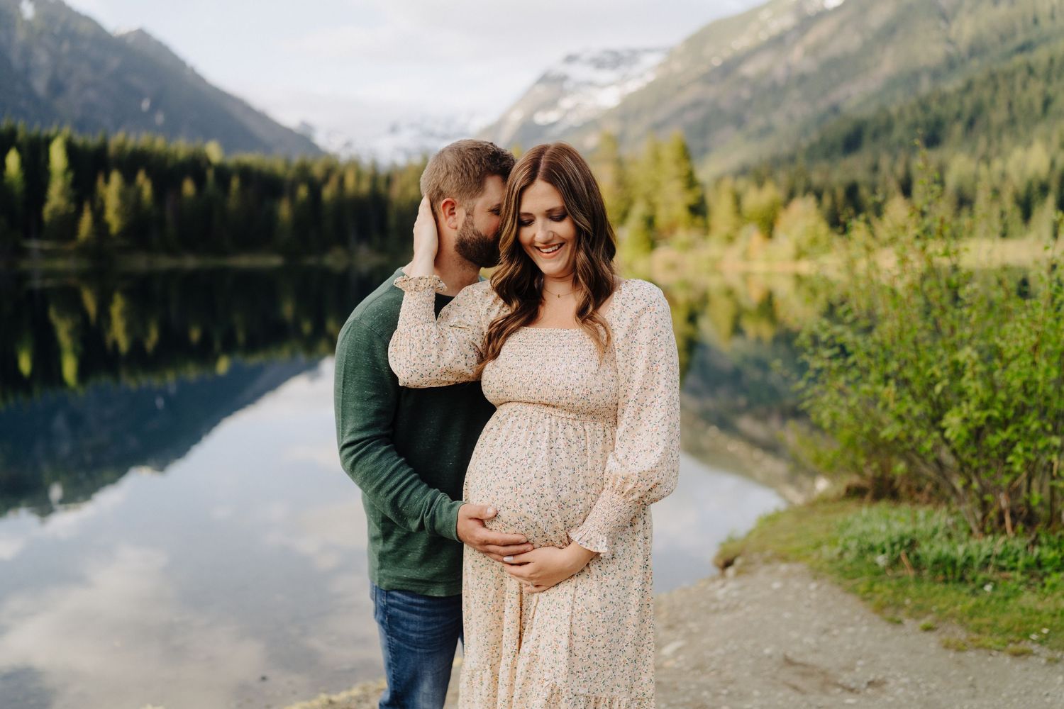 Couple poses together near tranquil lake with mountain backdrop and autumn foliage.