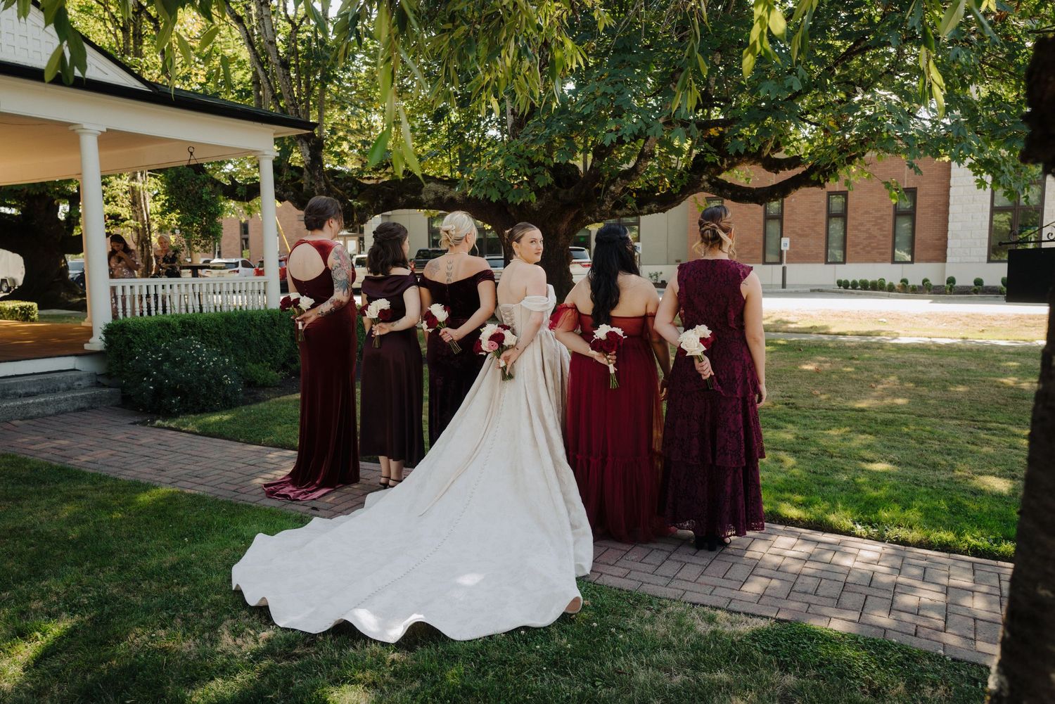 A bridal party in burgundy dresses poses outdoors on a garden path with trees in the background.