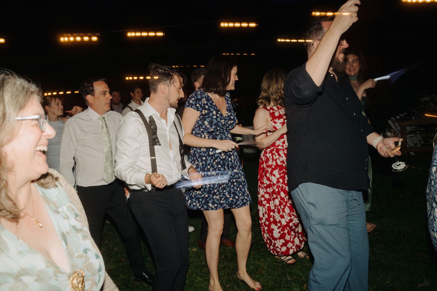Evening wedding reception with guests dancing under string lights in outdoor setting.