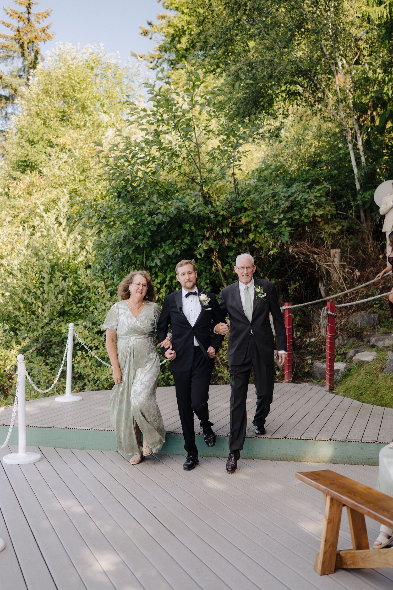 Wedding party members walk down an outdoor wooden deck pathway surrounded by lush greenery.