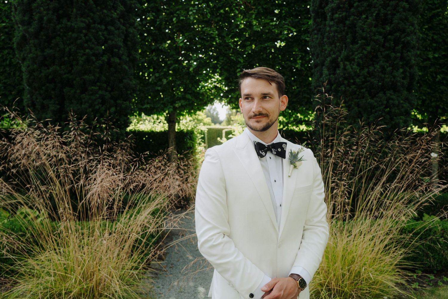 Groom in white tuxedo jacket and black bowtie stands in garden setting.