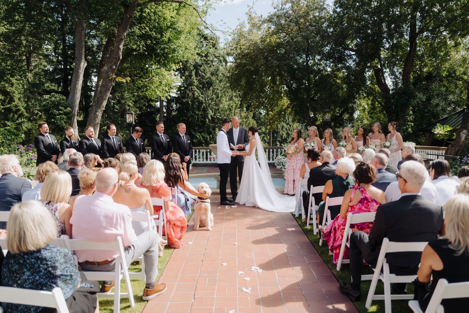 Wedding guests seated in white chairs watch ceremony on a brick pathway surrounded by trees.
