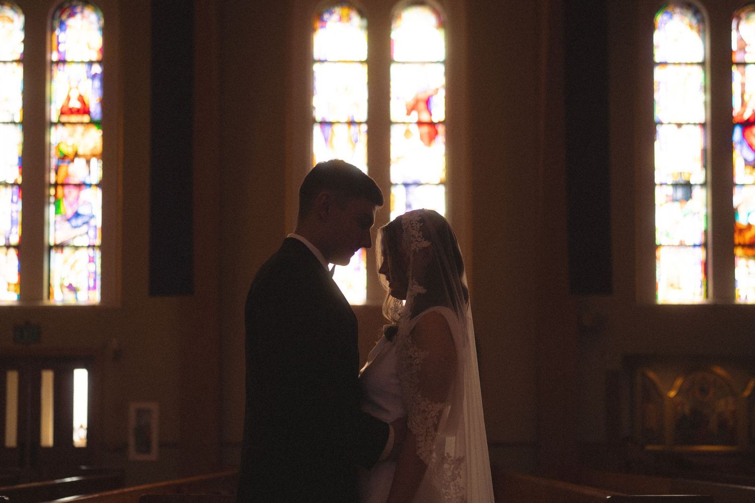 Bride and groom sharing an intimate moment in a dimly lit church with colorful stained glass windows behind them.