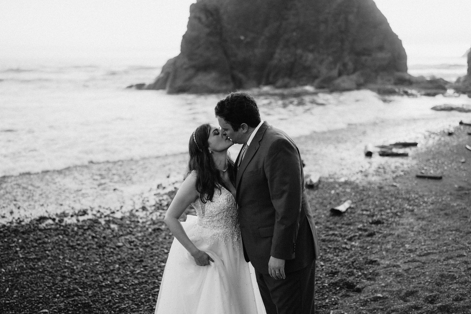 A black and white beach wedding photo with dramatic coastal rocks in the background.