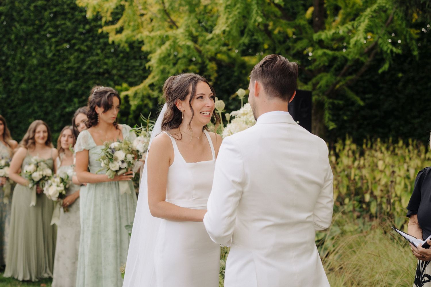 Garden wedding ceremony with bridesmaids in sage green dresses and white floral bouquets.