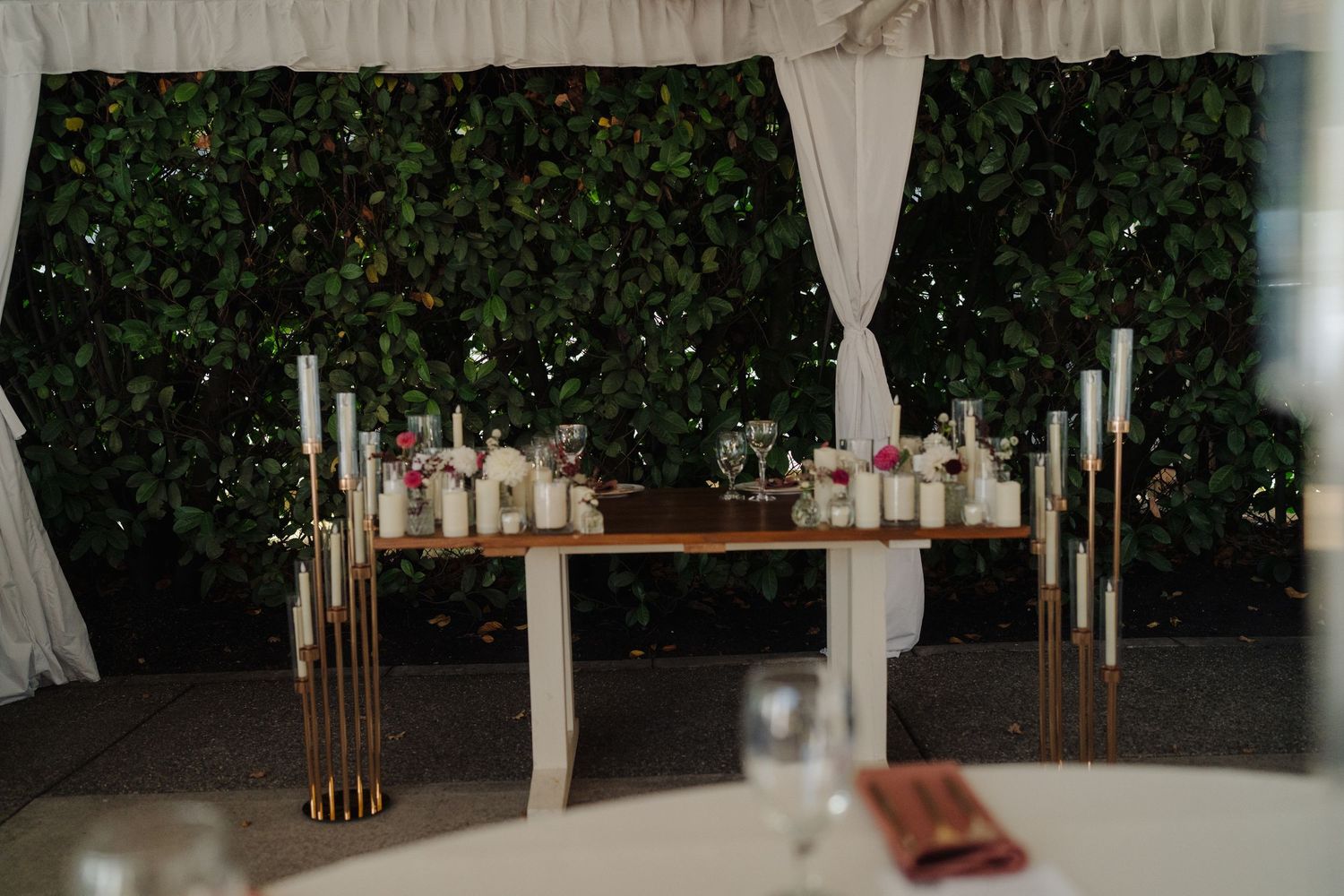 Wedding reception setup with wooden tables and greenery backdrop at outdoor venue.