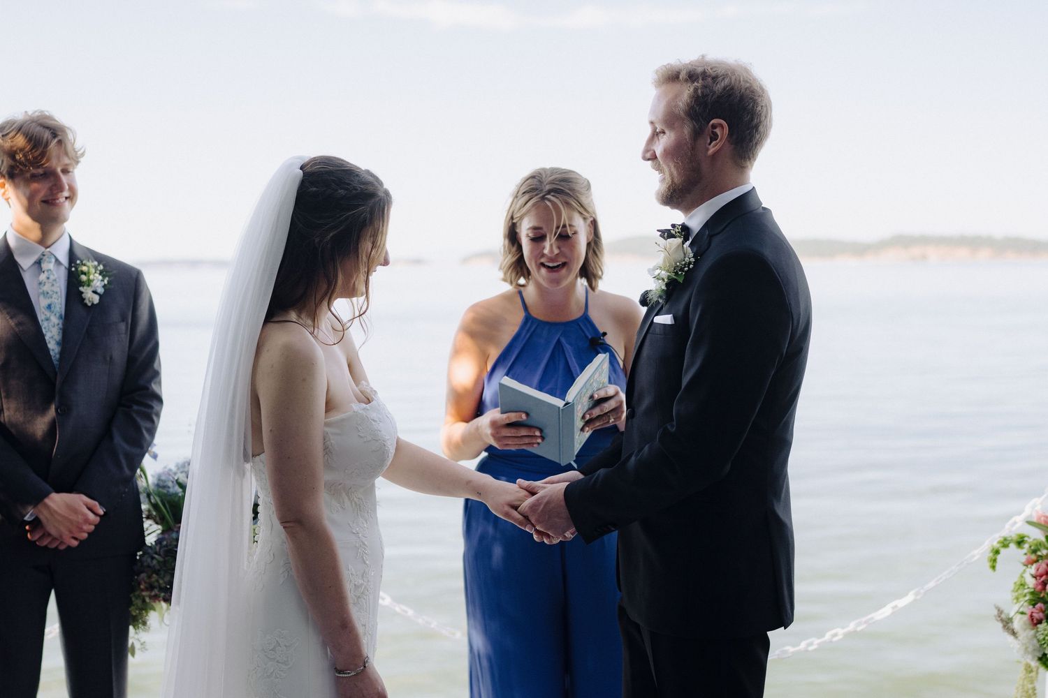 Seaside wedding ceremony with couple exchanging vows led by officiant in royal blue dress.