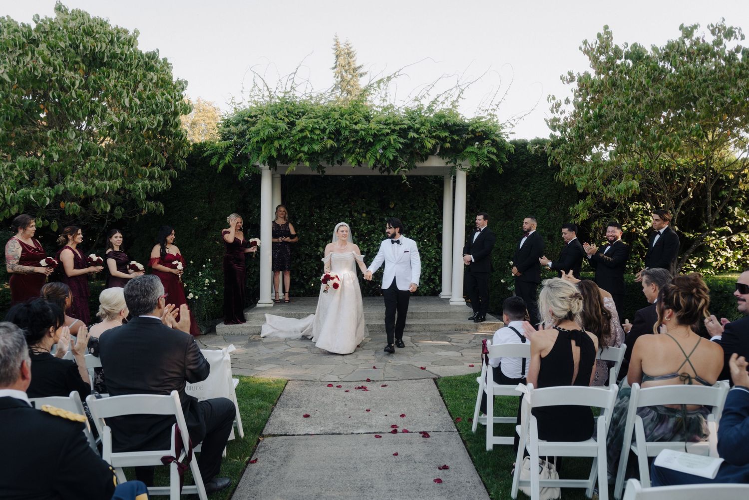 Outdoor wedding ceremony with guests watching couple exchange vows under a white arbor.