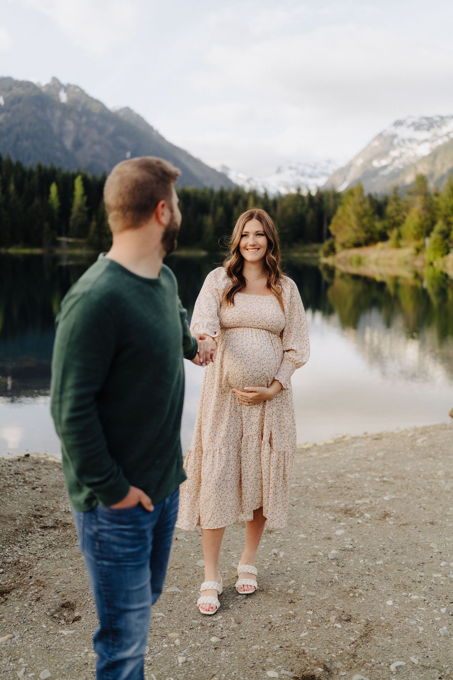A couple shares a tender moment by a serene mountain lake surrounded by evergreen trees and snowy peaks.