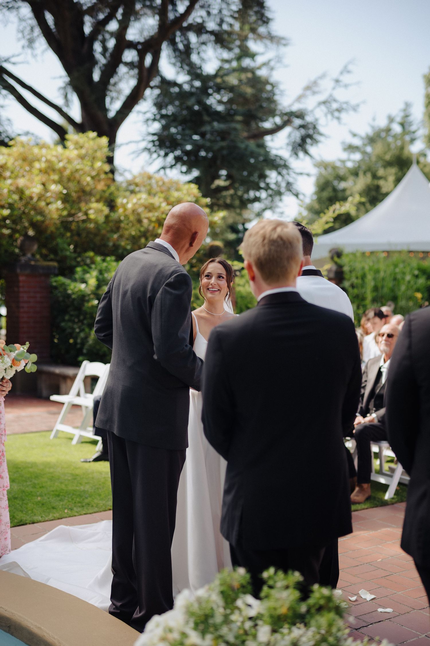 Wedding ceremony taking place outdoors with guests seated on white chairs and floral decorations.