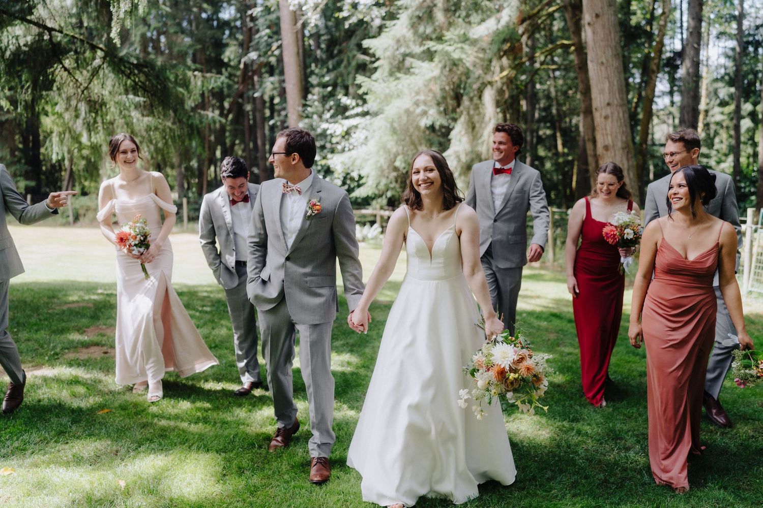 Wedding party walks through wooded path wearing burgundy dresses and gray suits.