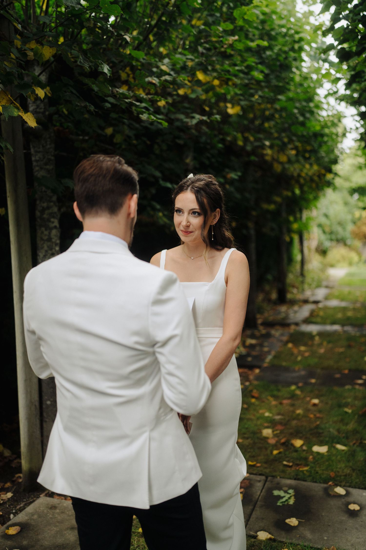 A couple in wedding attire share a tender moment together in a garden path lined with trees and fallen leaves.
