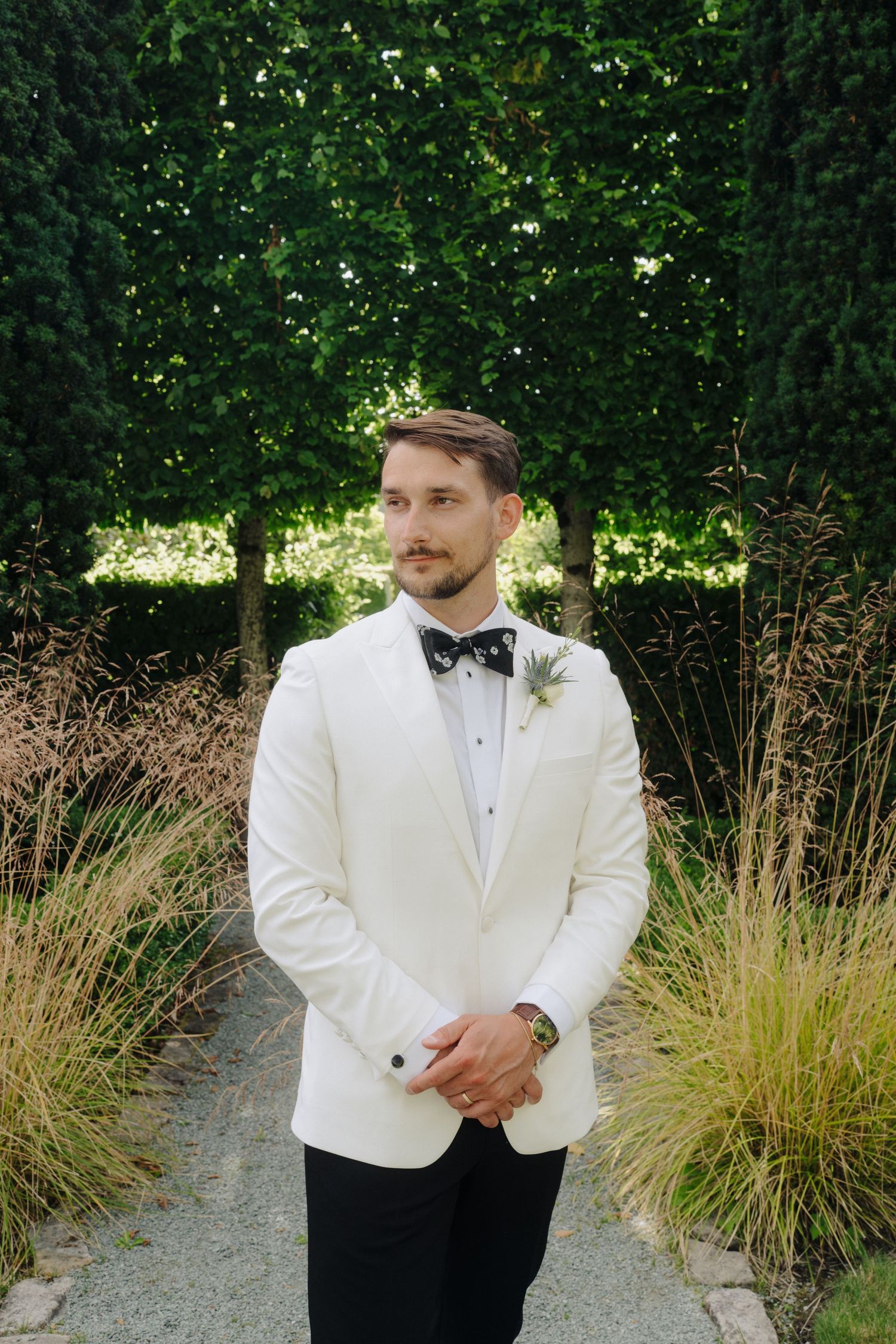 A groom in a classic white tuxedo jacket and black bow tie poses in front of lush greenery on his wedding day.