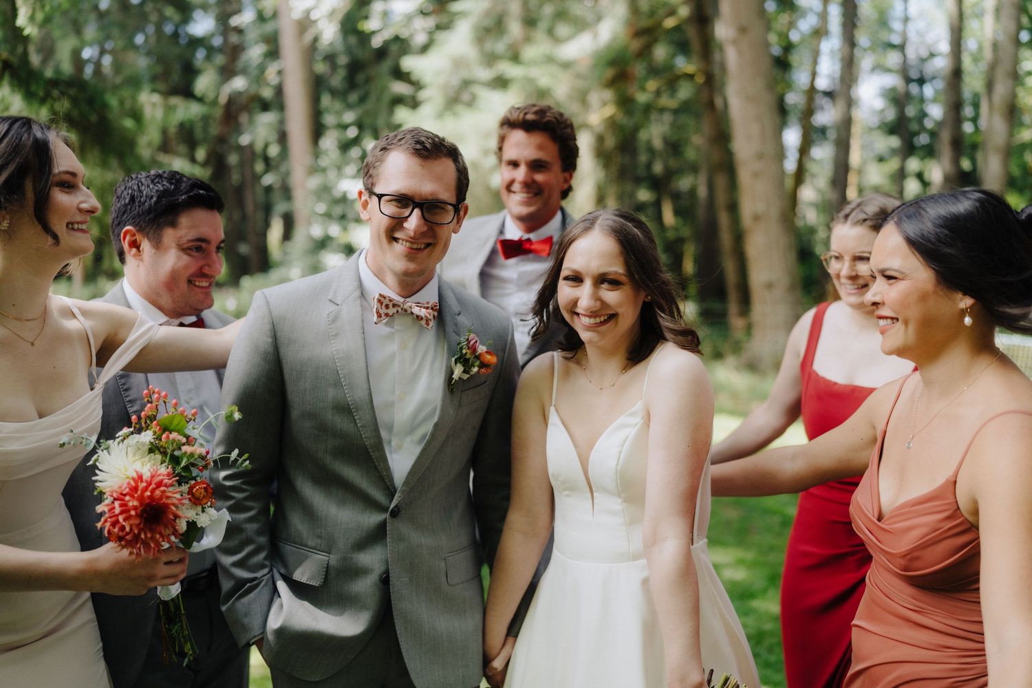 Wedding party celebrates joyfully outdoors with bridesmaids in burgundy dresses and groomsmen in gray suits.