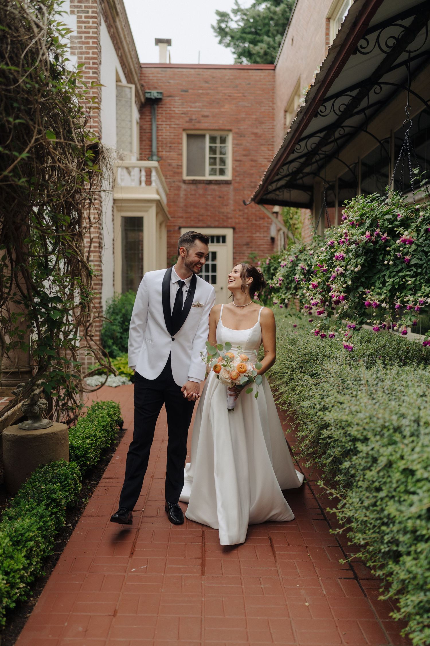 A couple in wedding attire poses in a garden pathway lined with brick walls and climbing roses.