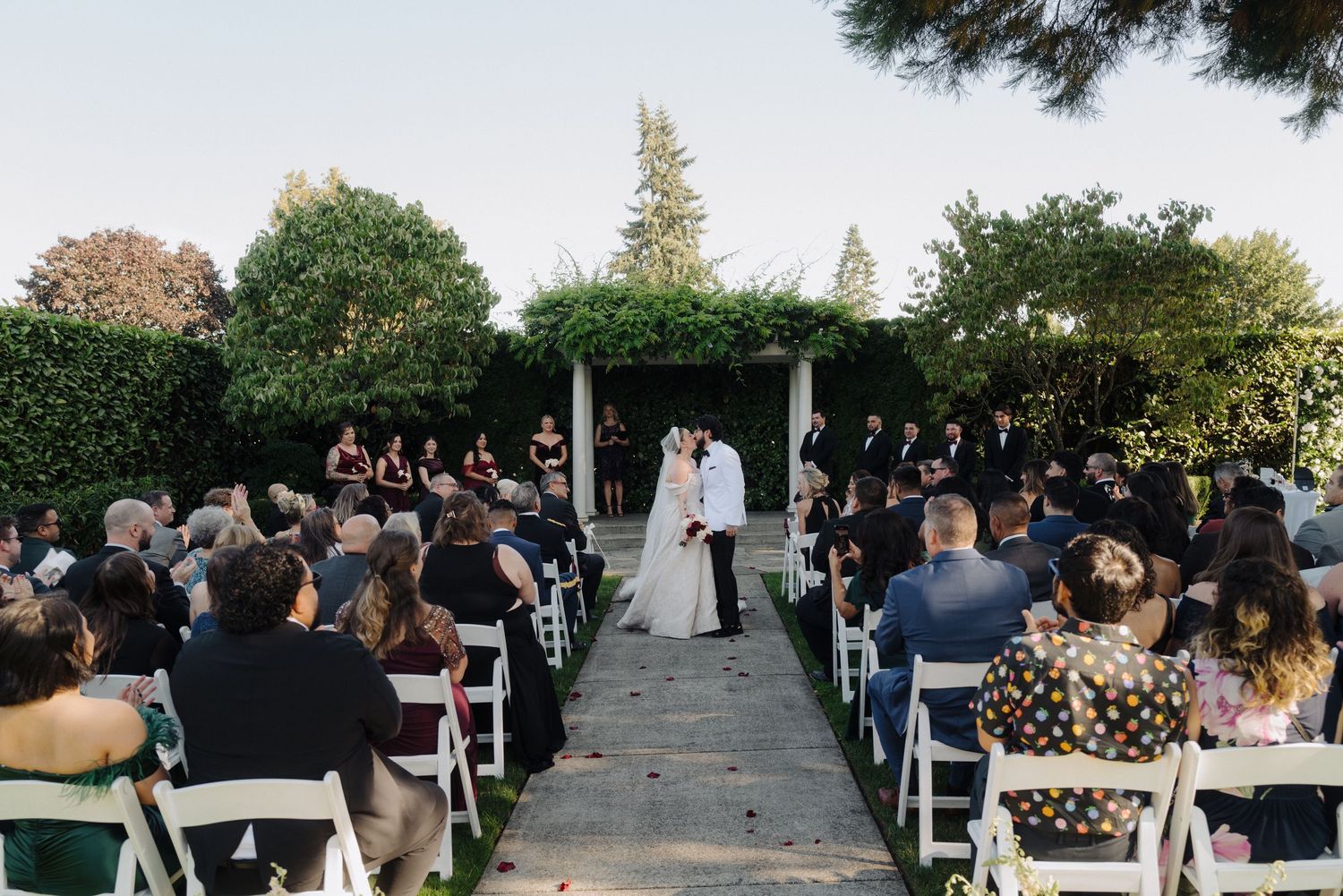 Panoramic view of a garden wedding ceremony with guests seated on white chairs.