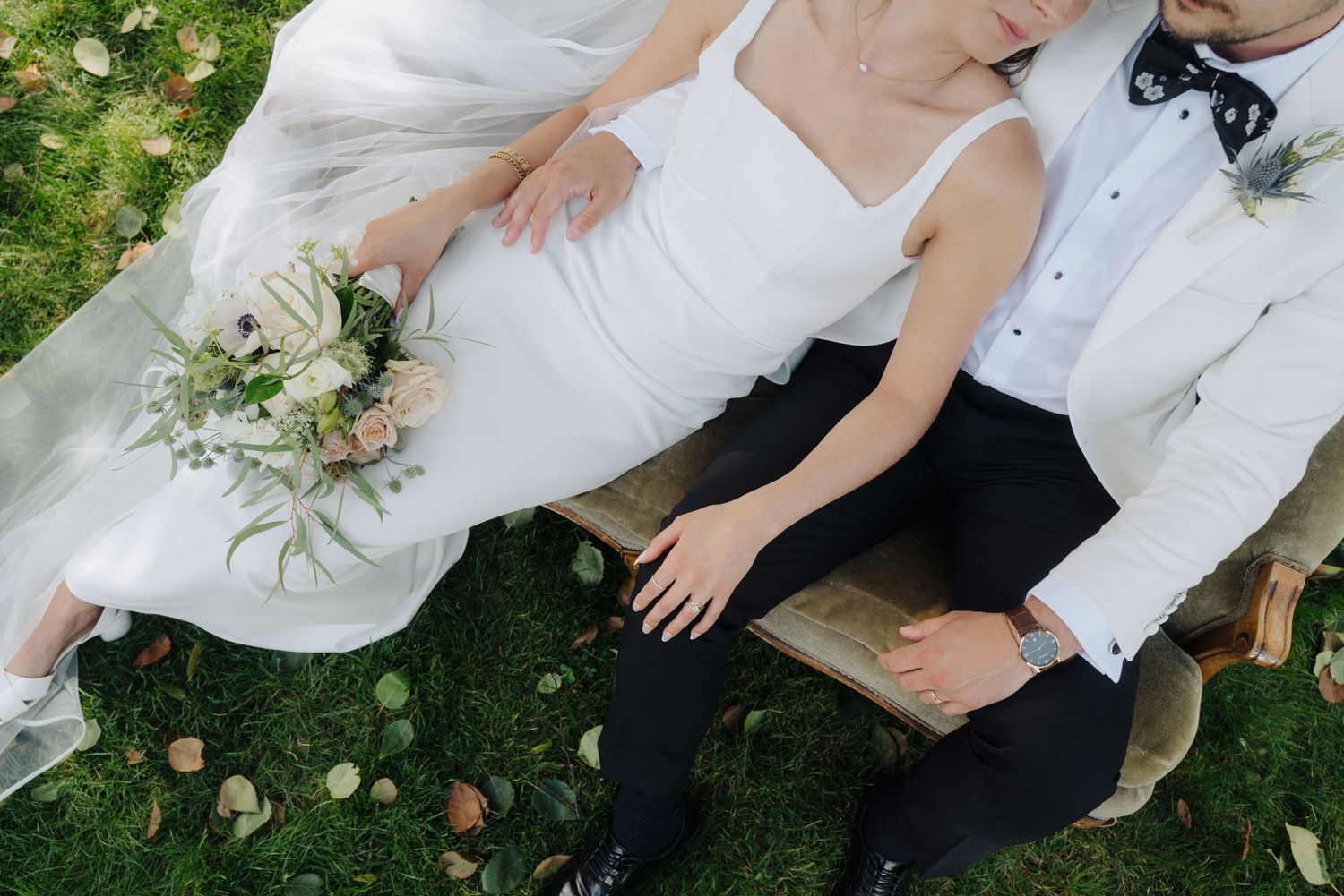 Couple in wedding attire sits together on grass with white floral bouquet.