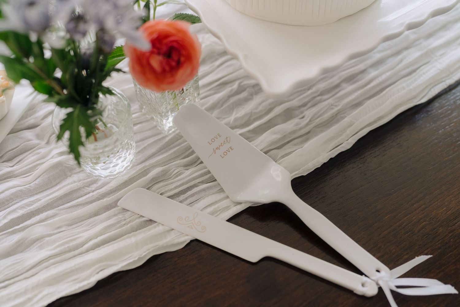 White wedding cake serving utensils with a coral rose on white linen tablecloth.