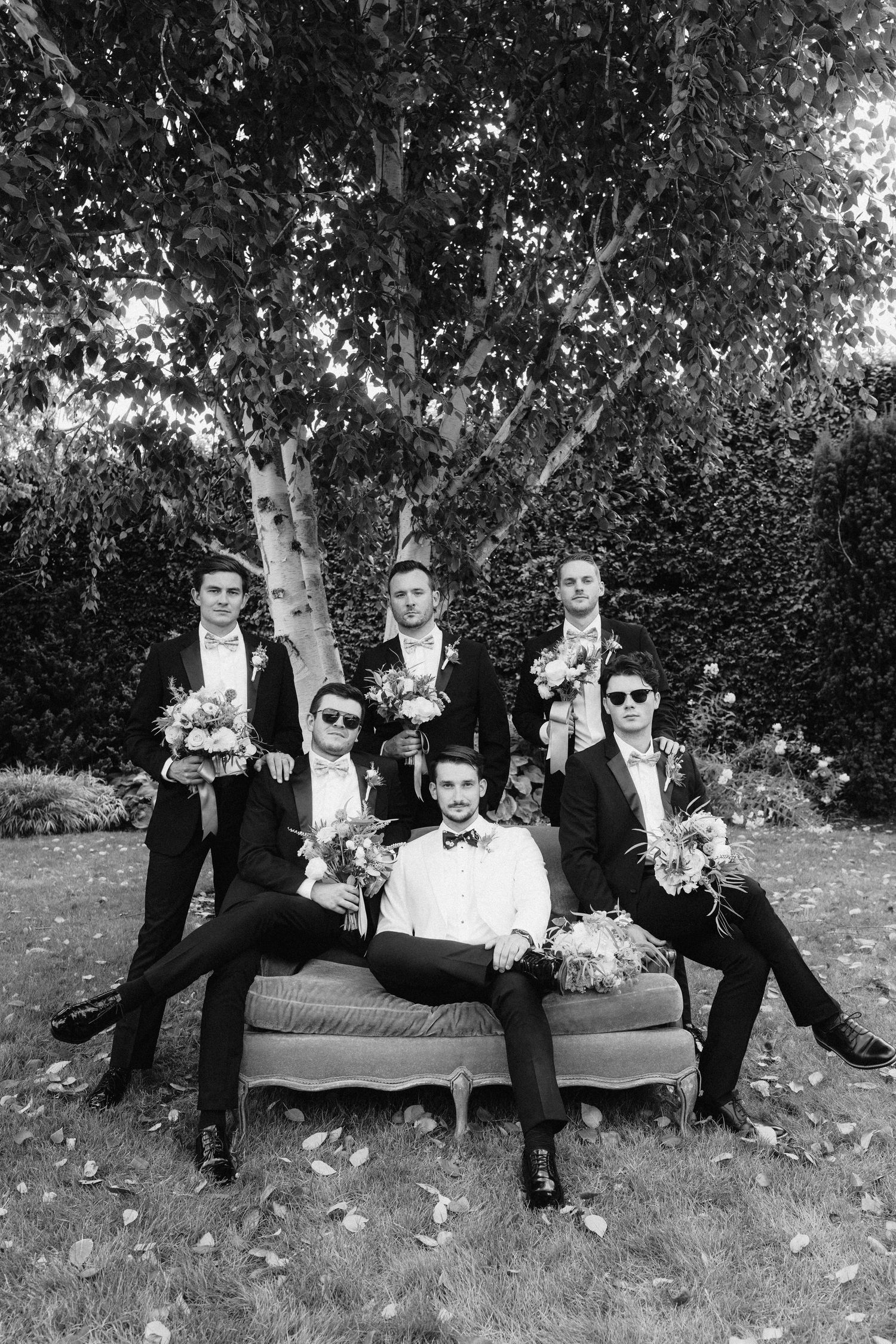 Groomsmen in dark suits lounging casually on grass during outdoor wedding photoshoot.