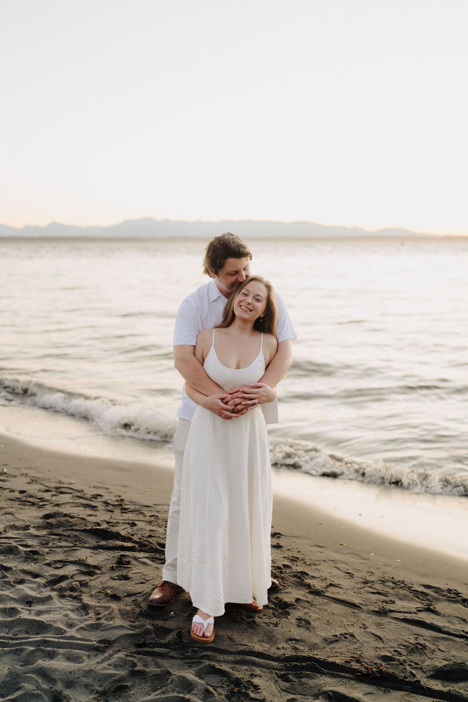 Intimate beach photoshoot featuring couple in white attire sitting on sandy shore at golden hour.