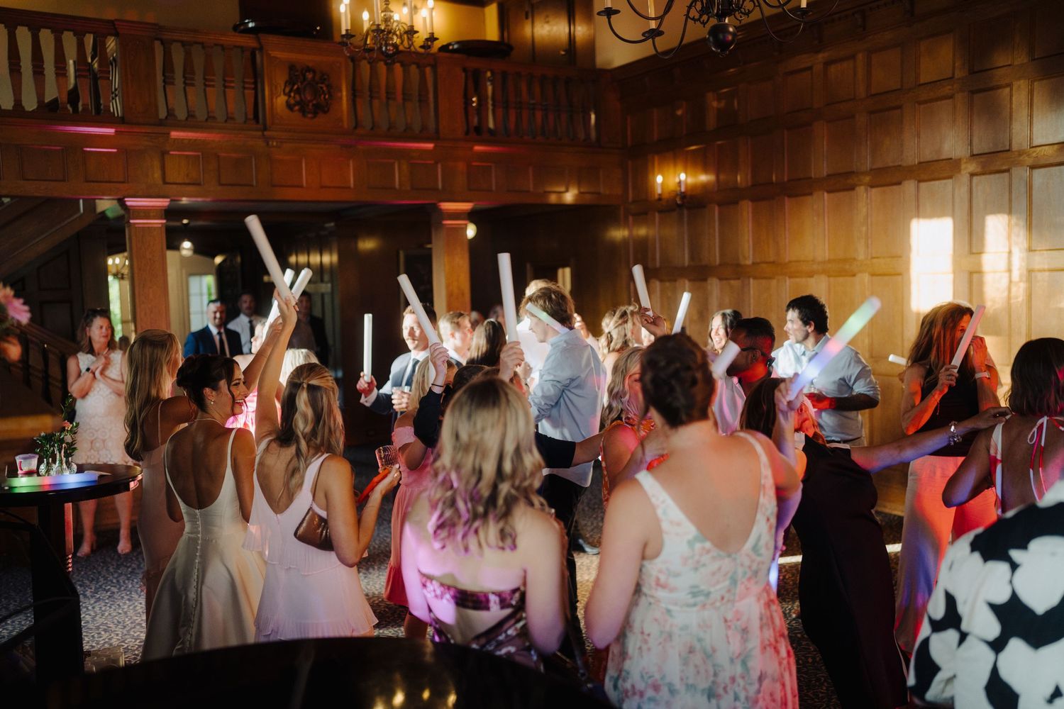 Wedding guests dancing and celebrating in an ornate wood-paneled ballroom with chandeliers.