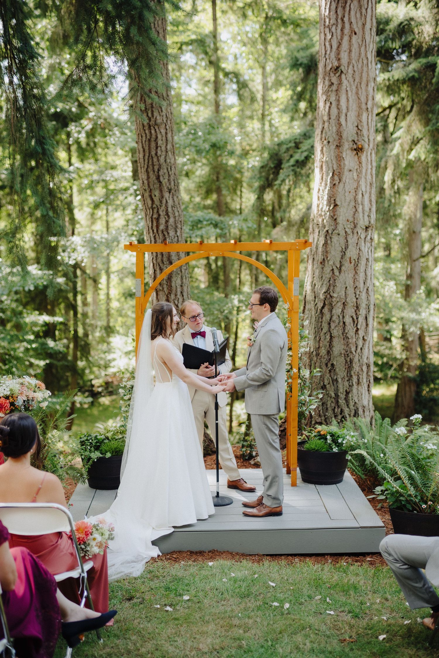An outdoor forest wedding ceremony under tall evergreen trees with a wooden platform and orange arch.