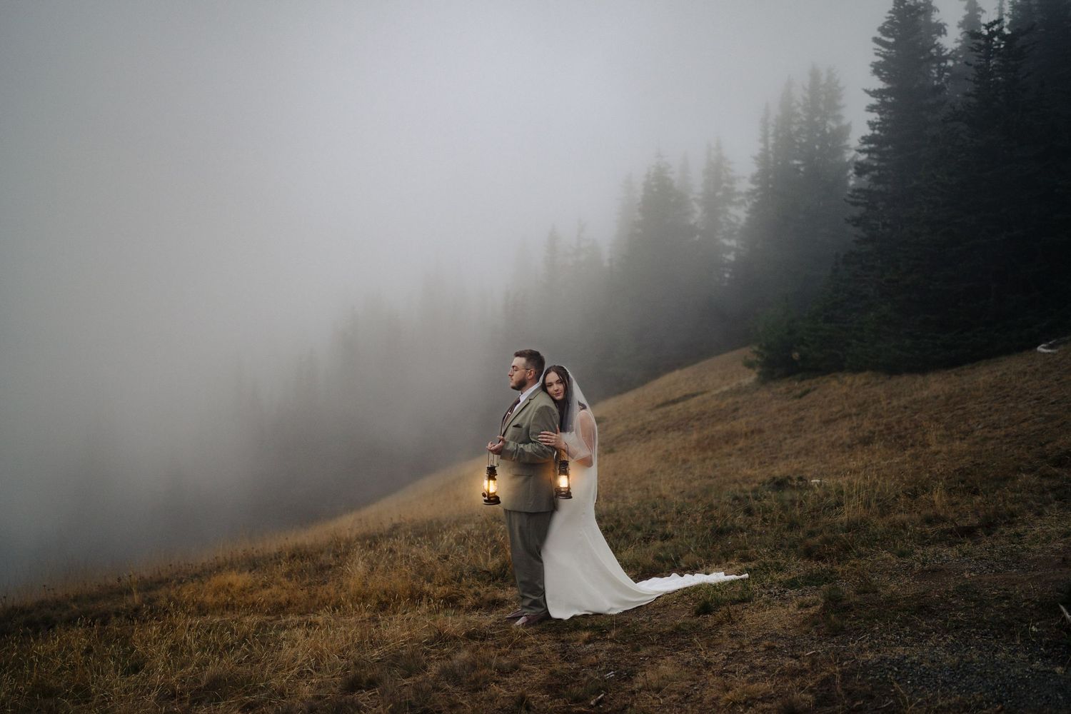 Wedding couple holding lanterns on a misty mountain hillside surrounded by fog and pine trees.