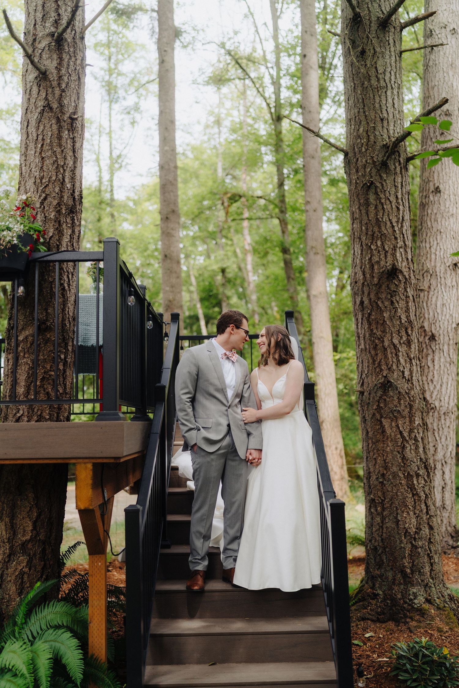 A couple in wedding attire stands together on wooden stairs surrounded by tall forest trees.