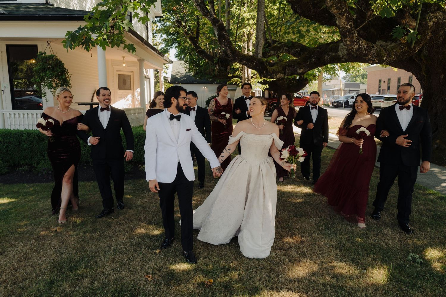 Wedding party in burgundy and formal attire poses casually on lawn in front of white house.