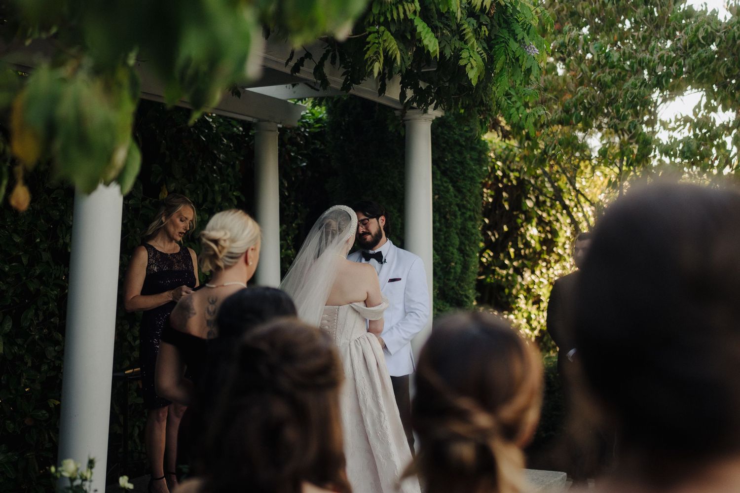 Wedding ceremony taking place under a white columned gazebo with guests watching during golden hour.