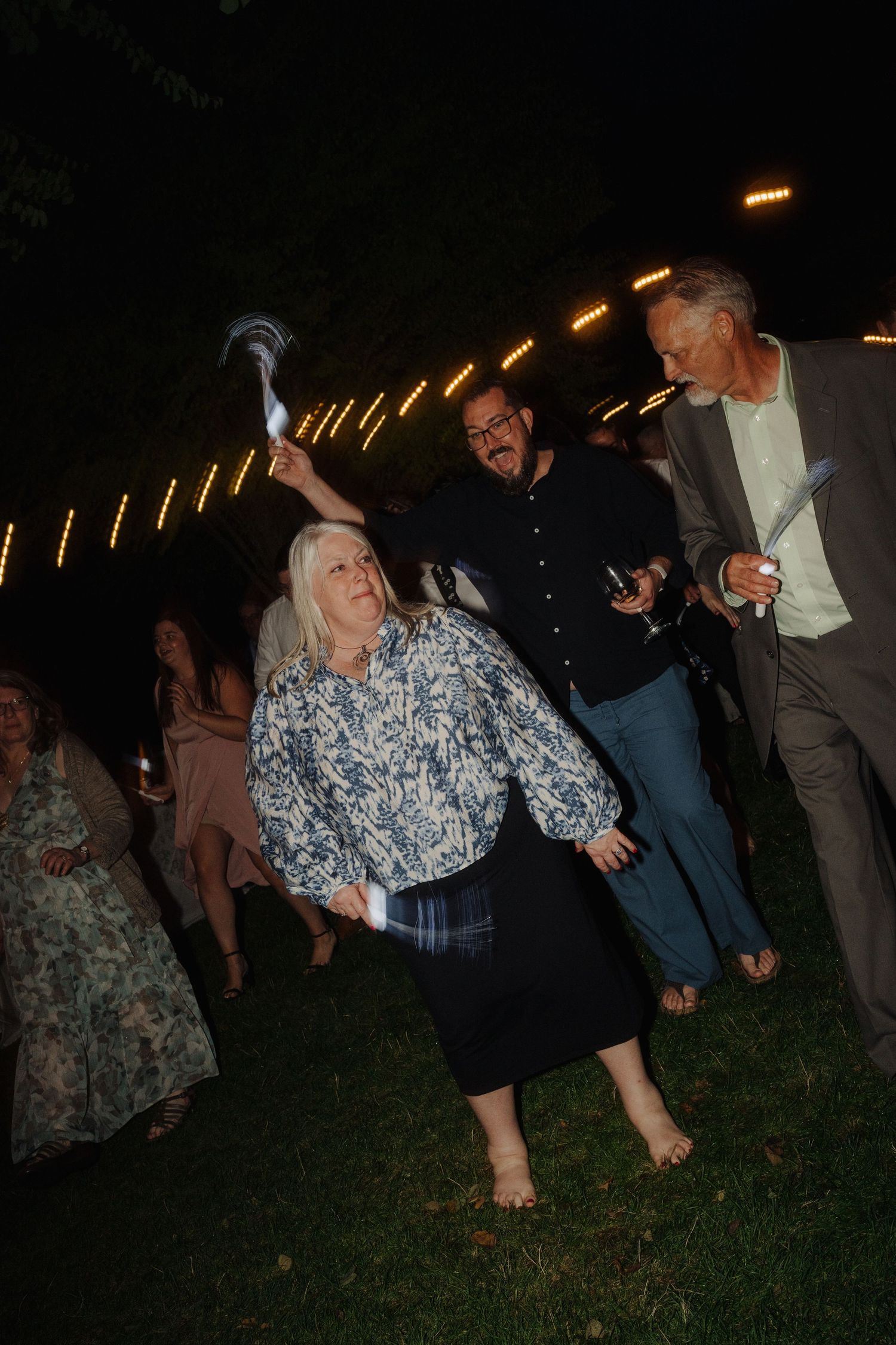 Party guests dance barefoot on the grass during a nighttime celebration.