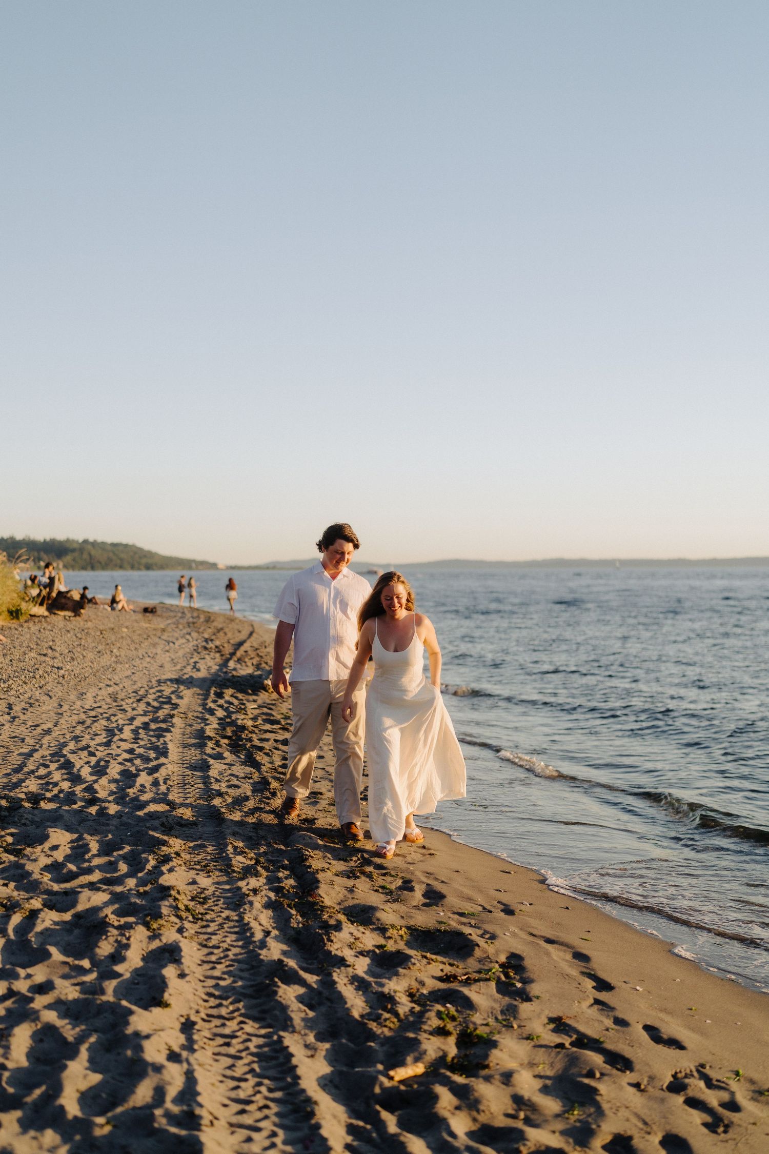 A couple walks hand in hand along a scenic beach shoreline during golden hour with waves lapping at the sand.