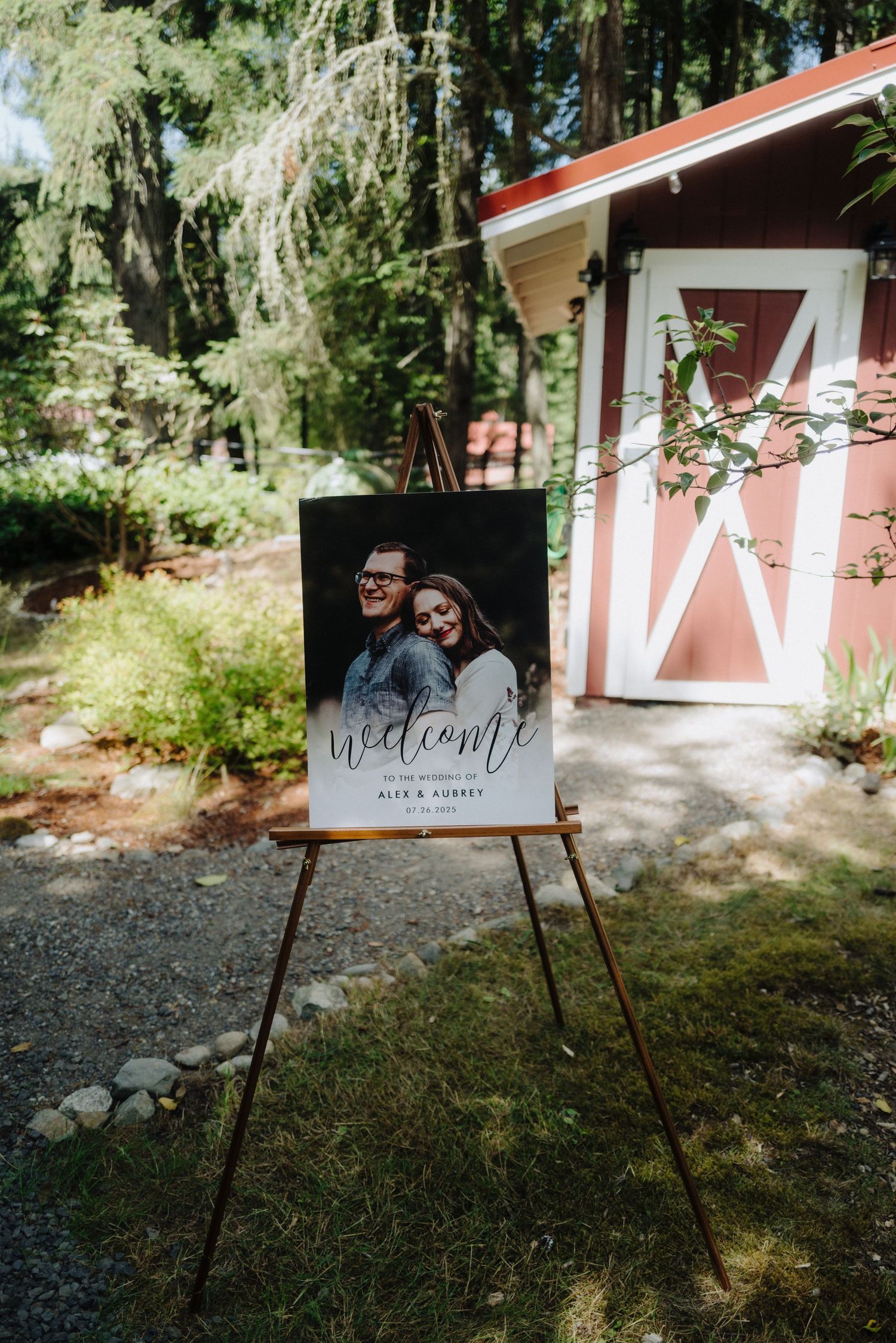 Wedding welcome sign displayed on easels outside a red barn venue in a wooded setting.