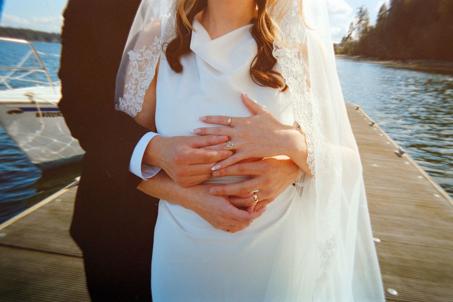 Bride and groom standing on a dock, hands clasped together showing wedding rings, with a lake in the background.