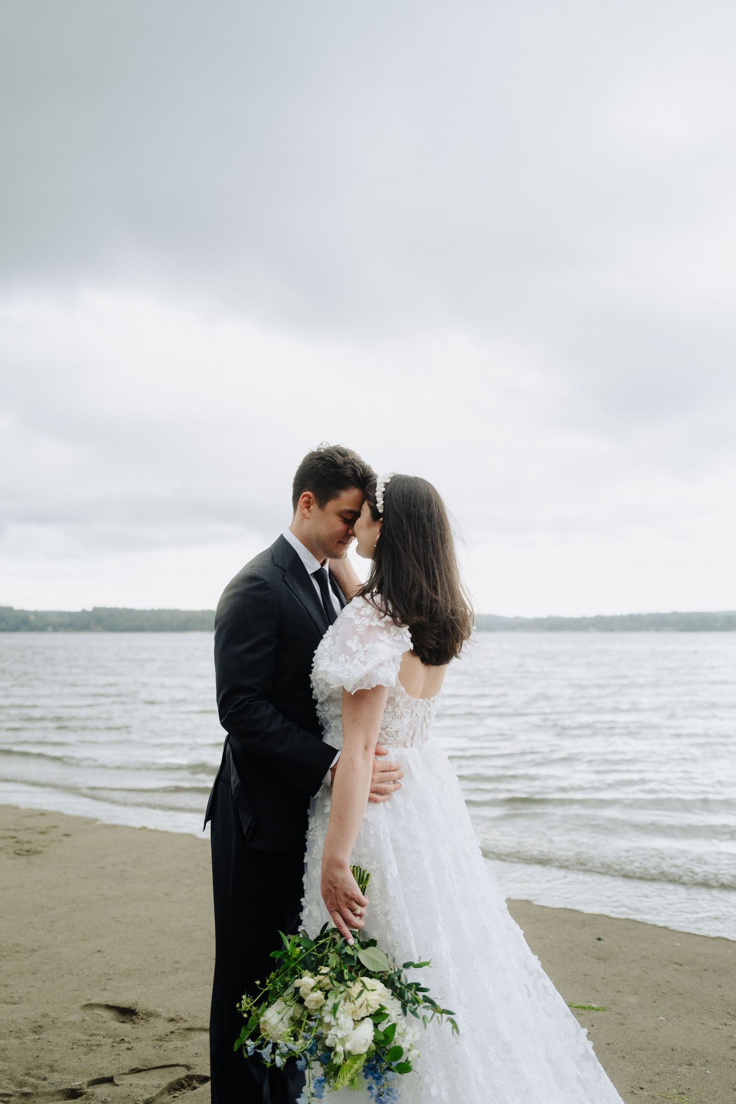 Romantic wedding moment captured on a cloudy beach with white floral bouquet.