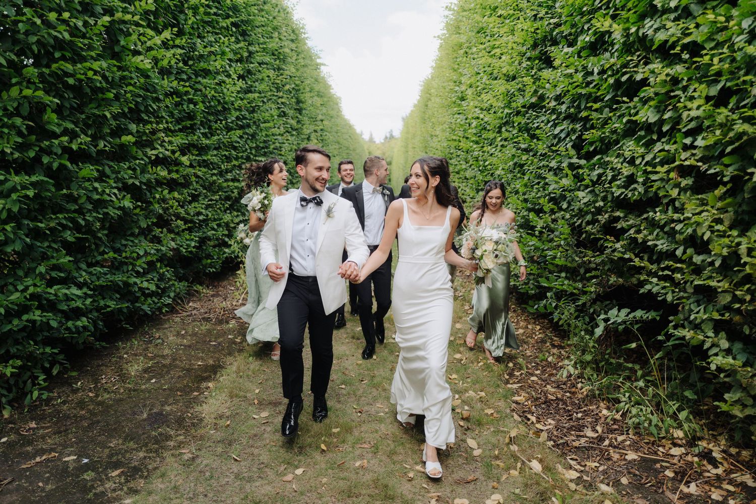 Wedding party walking through a lush green garden pathway lined with tall hedges.
