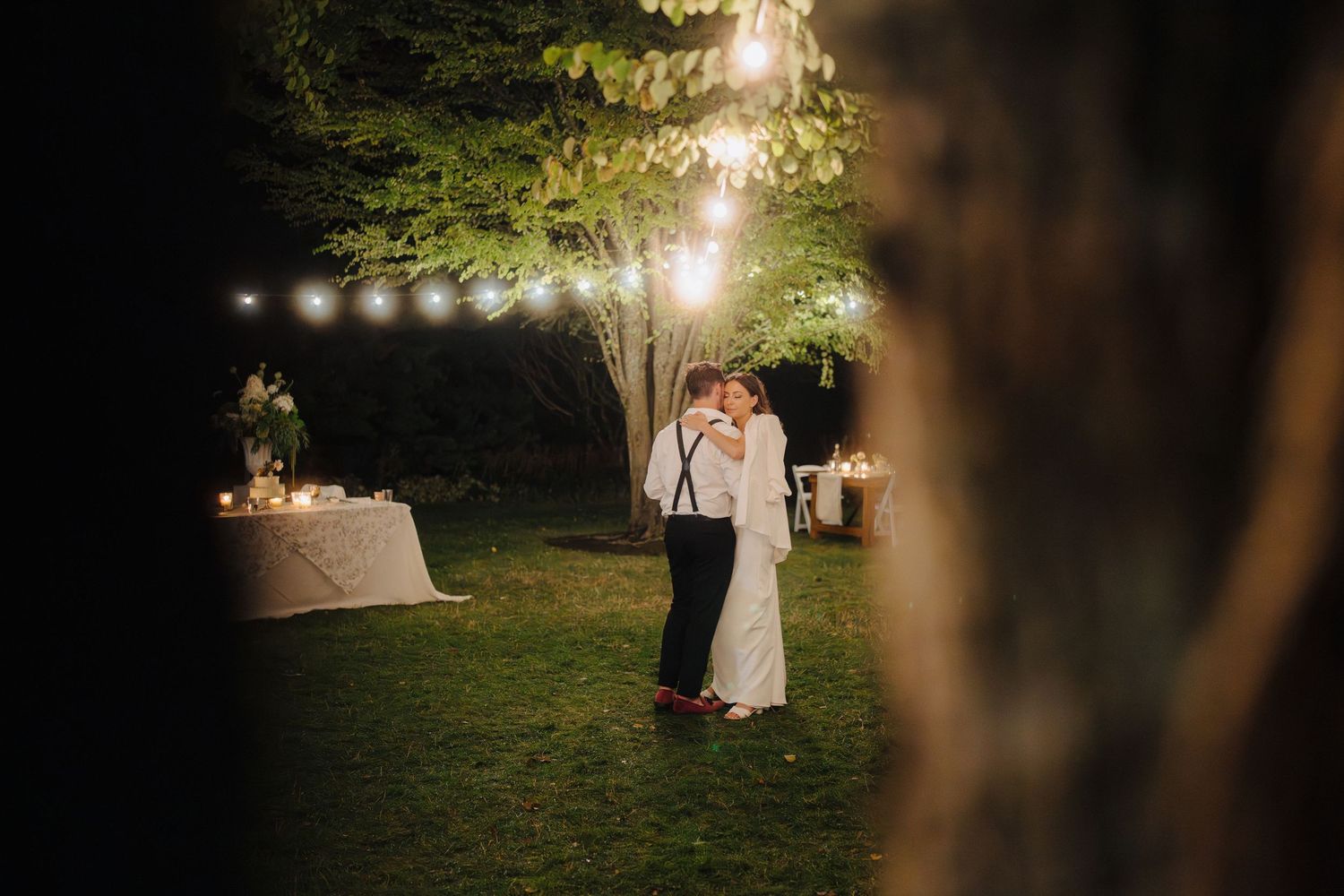 Romantic evening wedding portrait under glowing string lights in garden setting.