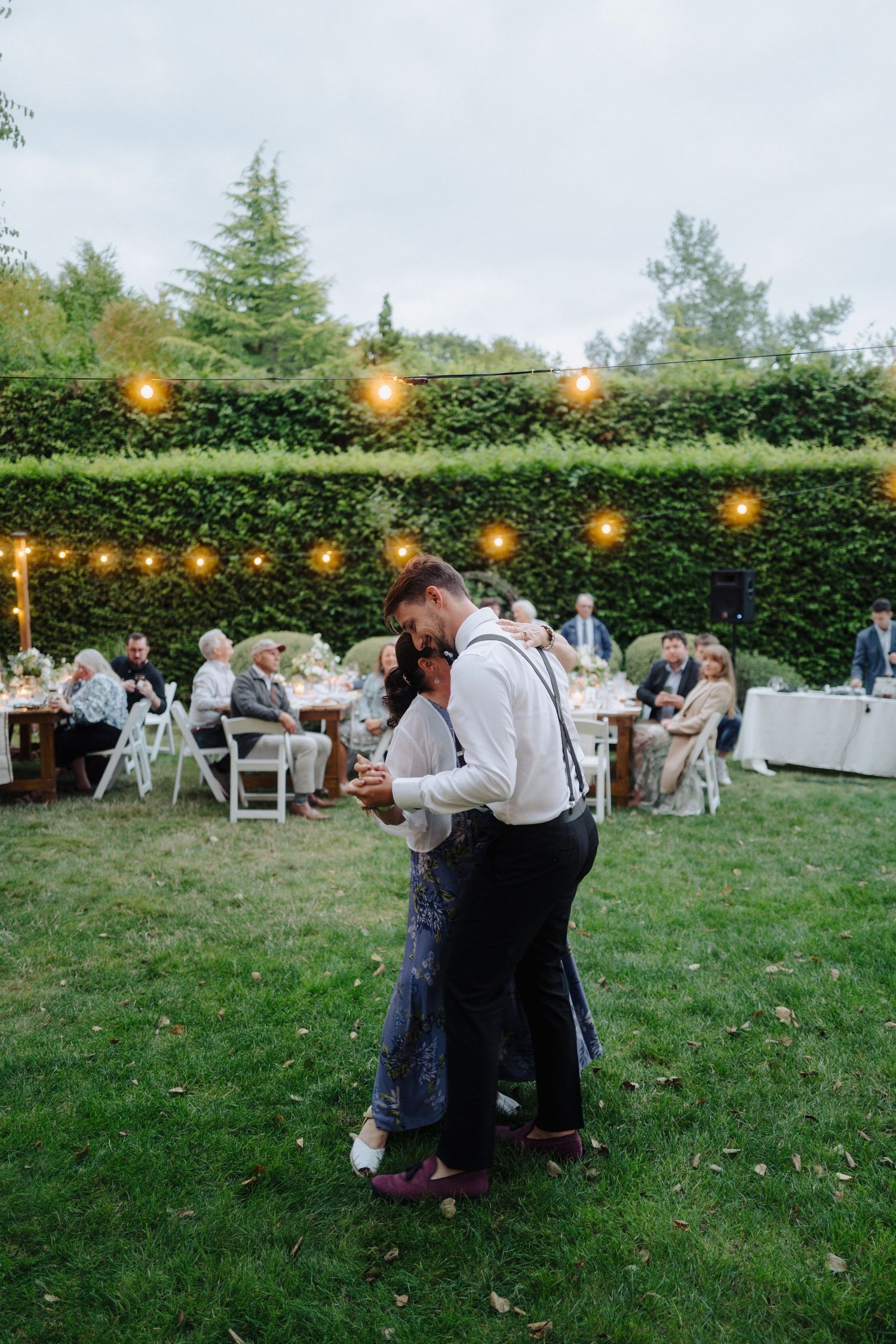 Guests dancing at a garden wedding reception with string lights illuminating the hedge backdrop.