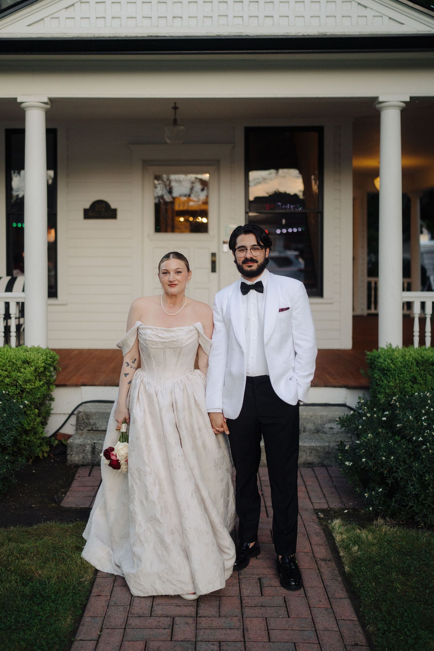 Newlyweds walking together on brick pathway in front of white colonial style house.