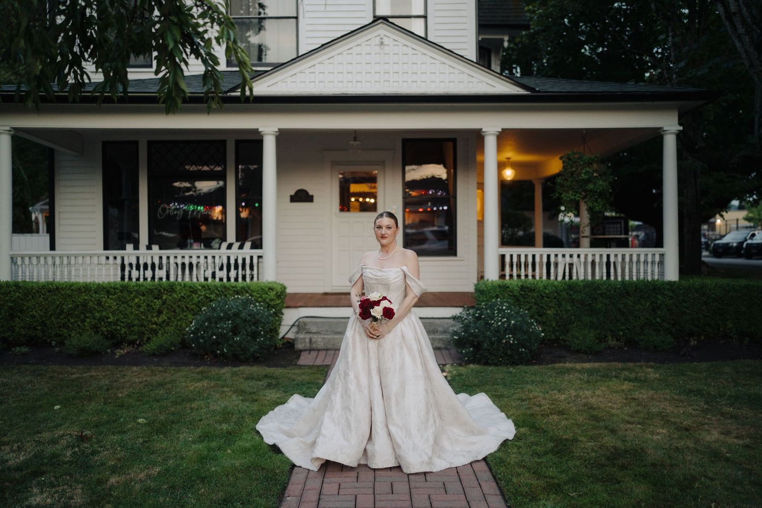 A wedding party poses on the front porch of a charming white house with elegant columns and landscaping.