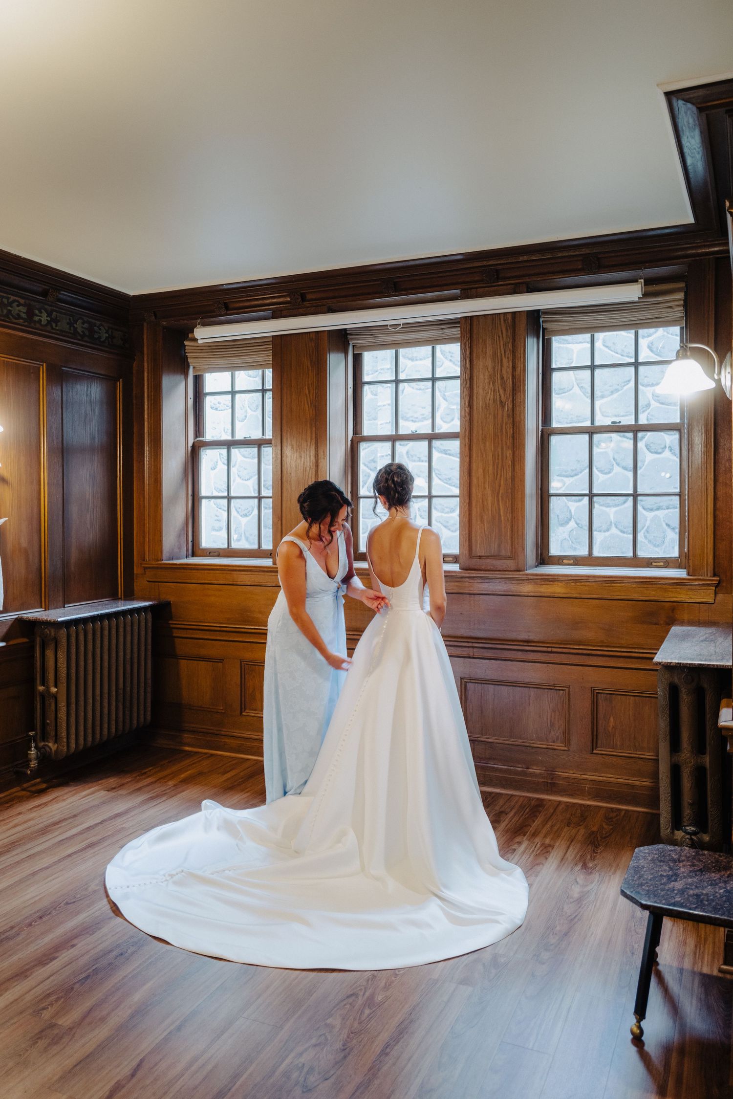 Elegant wood-paneled room with large windows showcases a flowing white wedding dress train.