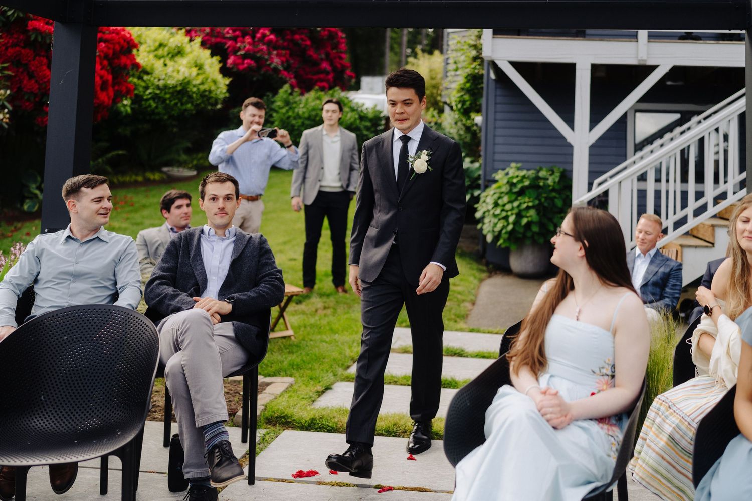 Wedding guests gather on a lush green lawn outside a white-trimmed building during an outdoor ceremony.