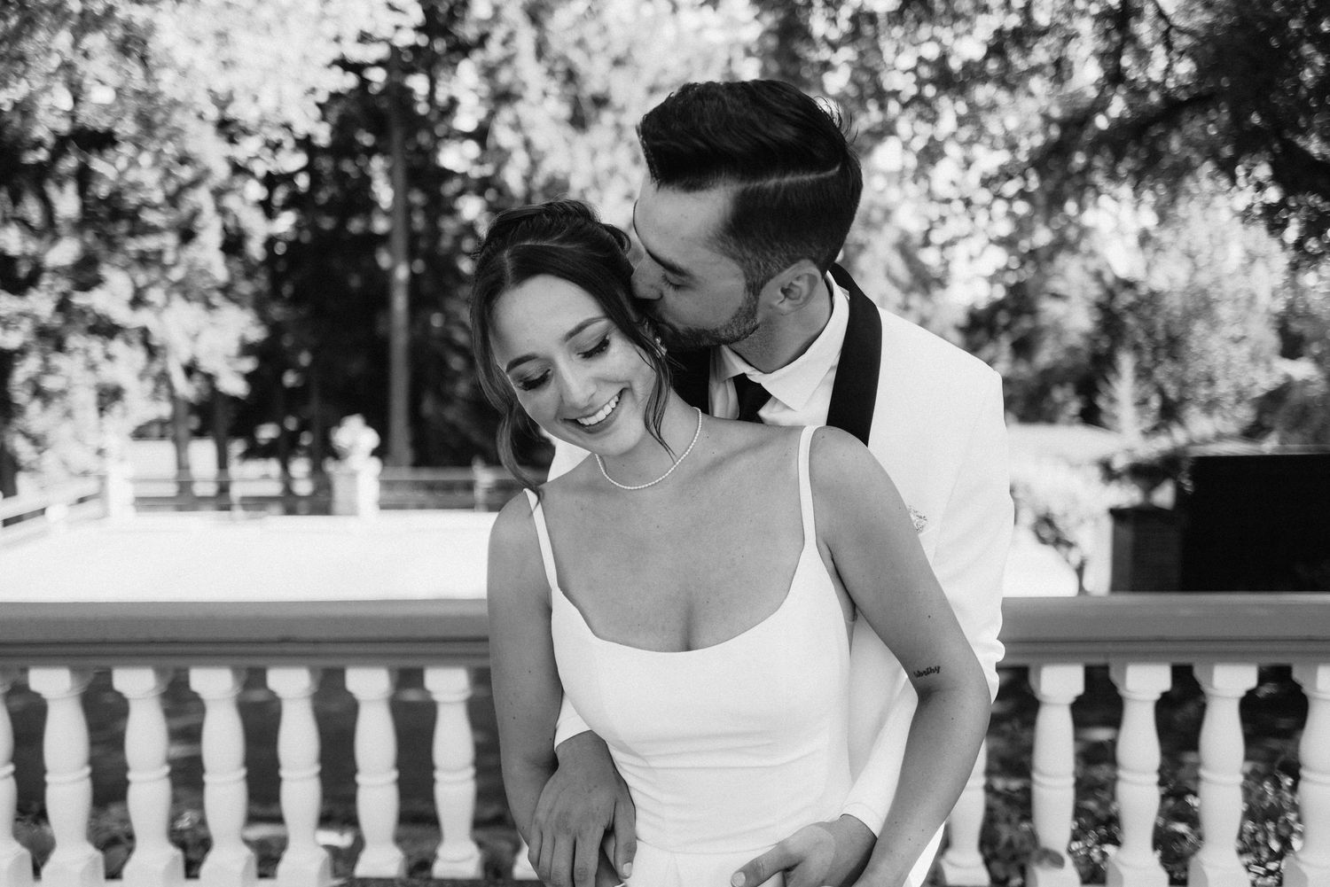 A black and white portrait of a couple sharing a tender moment on a balcony with stone railings.