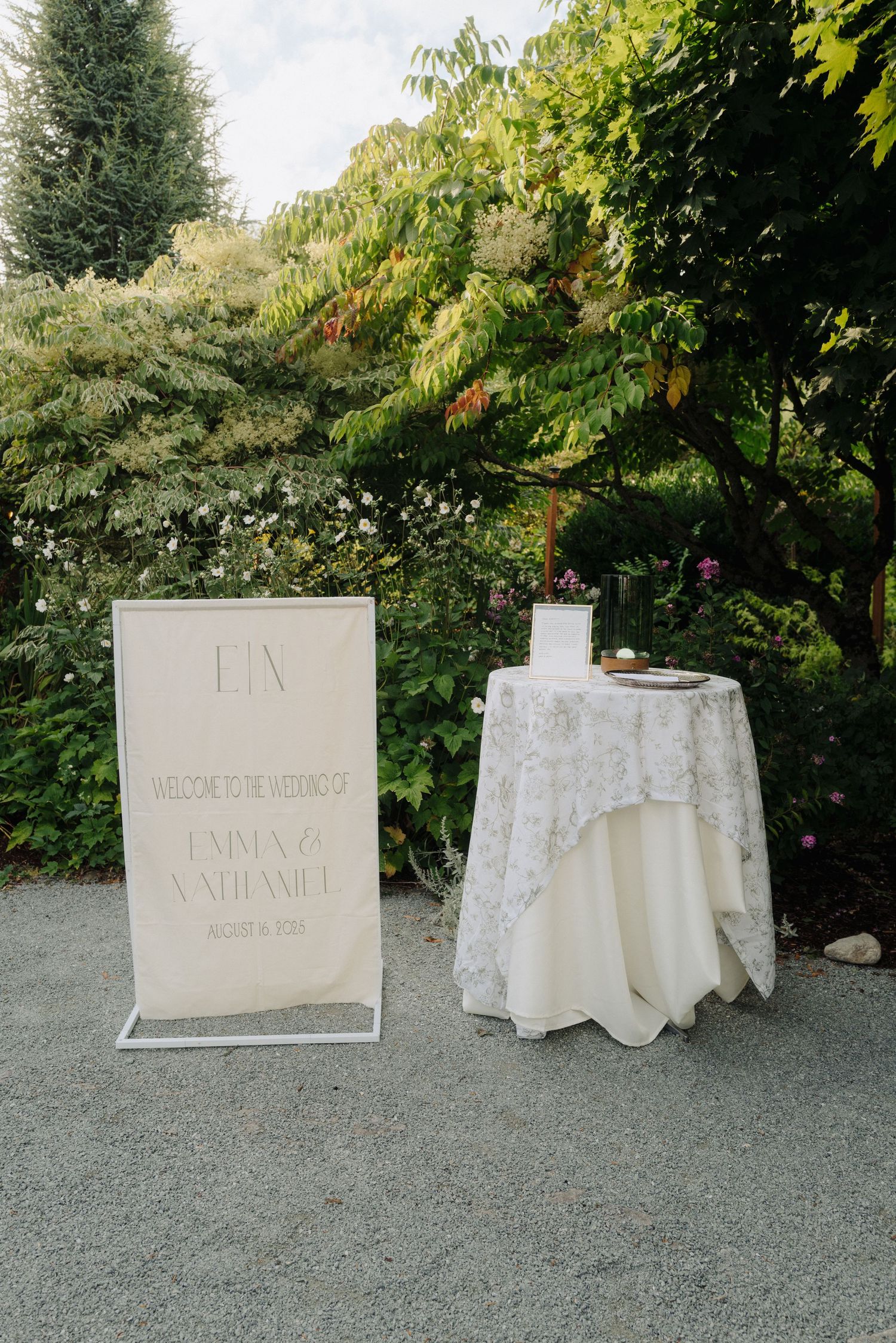 Wedding reception setup in a garden with a white tablecloth and signage against lush greenery.