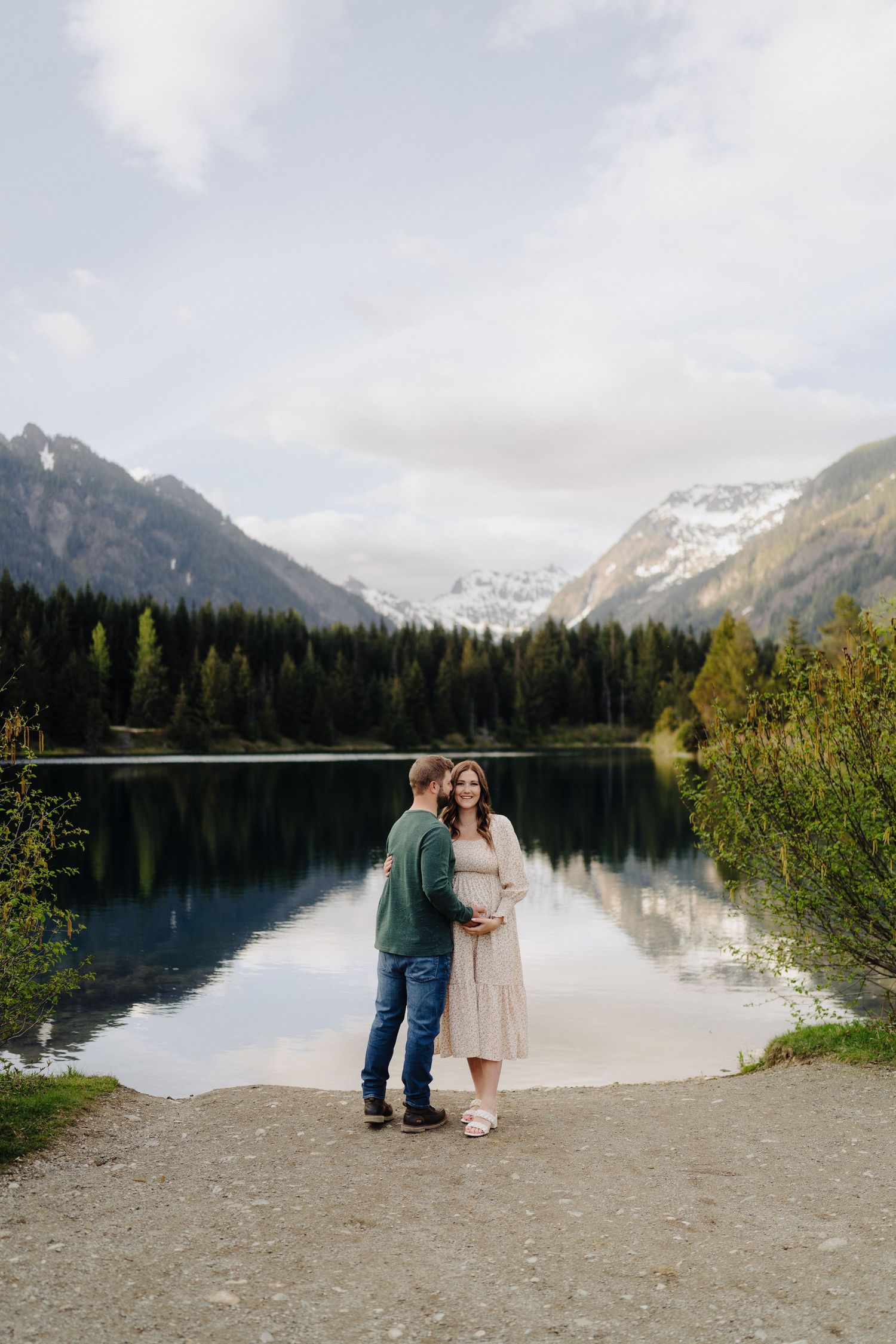 A couple shares an intimate moment by a serene mountain lake surrounded by snow-capped peaks and evergreen forest.